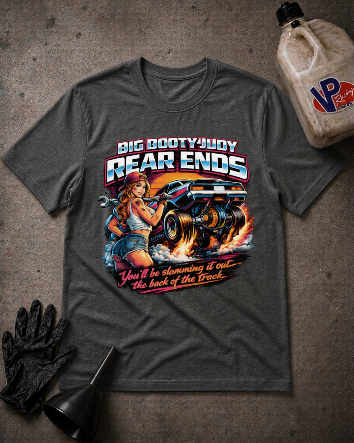 A dark gray graphic t-shirt laid flat on a textured gray surface, surrounded by racing-themed props including a black oil funnel, a black glove, and a plastic jug of VP Racing. The t-shirt features a large graphic of a pin-up girl and a monster truck with the text 'BIG BOOTYJUDY REAR ENDS' and 'You'll be slamming it out the back of the Track'.