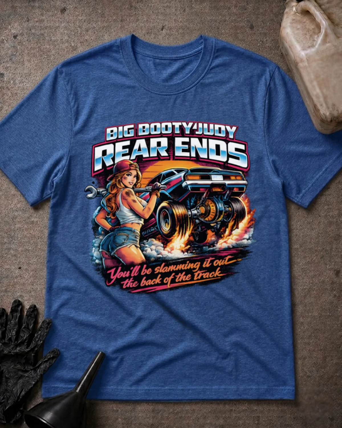A heather true royal  graphic t-shirt laid flat on a textured gray surface with automotive props. The graphic features a pin-up style illustration of a woman holding a wrench next to a race car, with the text 'BIG BOOTYJUDY REAR ENDS' and 'You'll be slamming it out the back of the Track'.