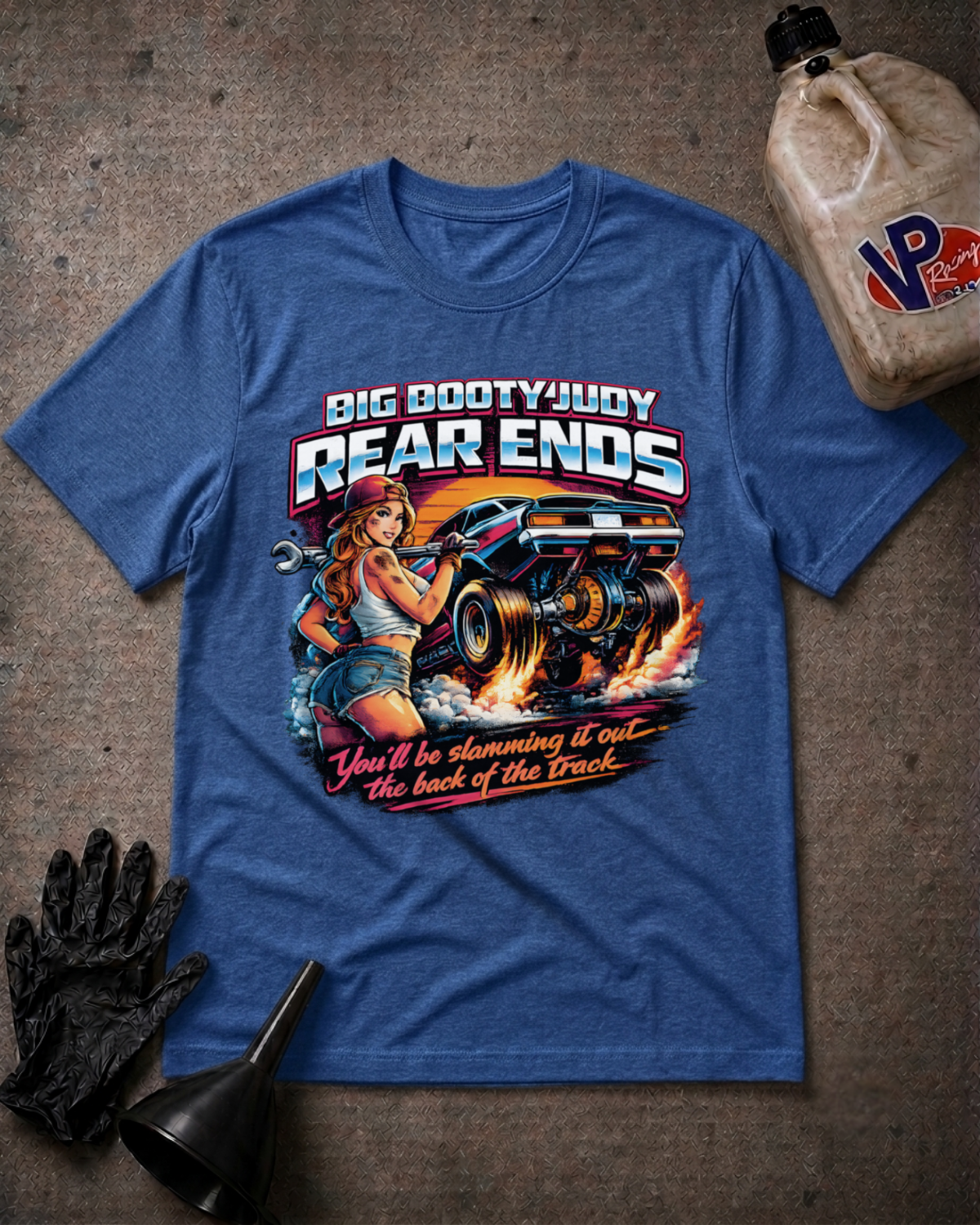 A heather true royal graphic t-shirt laid flat on a textured gray surface, surrounded by racing-themed props including a black oil funnel, a black glove, and a plastic jug of VP Racing. The t-shirt features a large graphic of a pin-up girl and a race car with the text 'BIG BOOTYJUDY REAR ENDS' and 'You'll be slamming it out the back of the Track'.