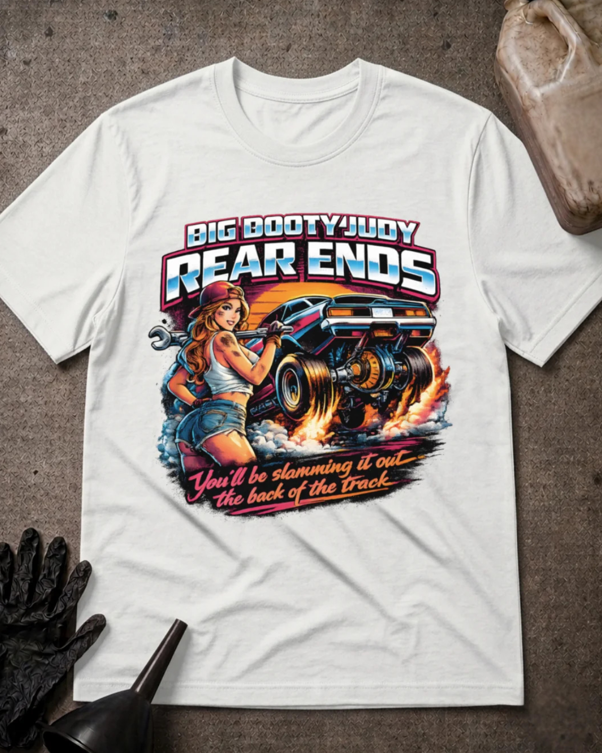 A white  graphic t-shirt laid flat on a textured gray surface with automotive props. The graphic features a pin-up style illustration of a woman holding a wrench next to a race car, with the text 'BIG BOOTYJUDY REAR ENDS' and 'You'll be slamming it out the back of the Track'.