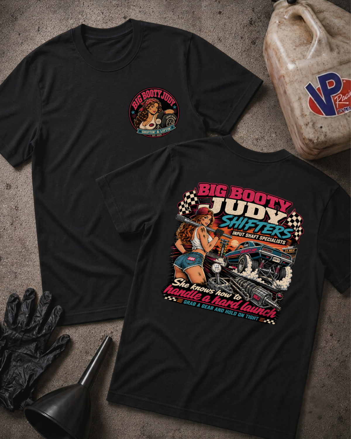 Two black graphic T-shirts laid flat on a textured gray surface, with racing-themed artwork and text including 'BIG BOOTY JUDY SHIFTERS' and 'She knows how to handle a hard launch.' Props around them include a plastic gas can, black gloves, a funnel, and a worn bucket.