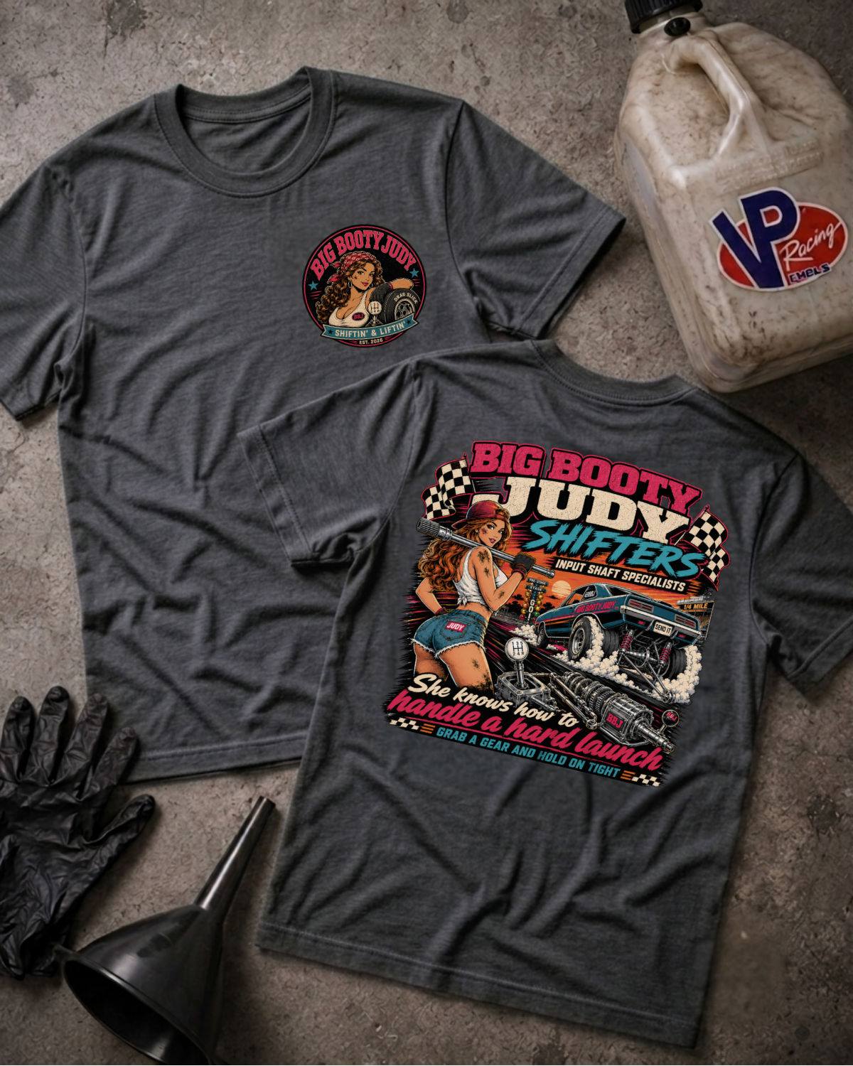 Two dark heather gray graphic t-shirts displayed flat on a textured concrete surface, accompanied by vintage-style garage props including a plastic fuel jug, black gloves, and a funnel. The front shirt features a small circular graphic on the left chest, while the back shirt displays a large retro-style graphic with the text 'BIG BOOTY JUDY SHIFTERS' and a pin-up illustration.