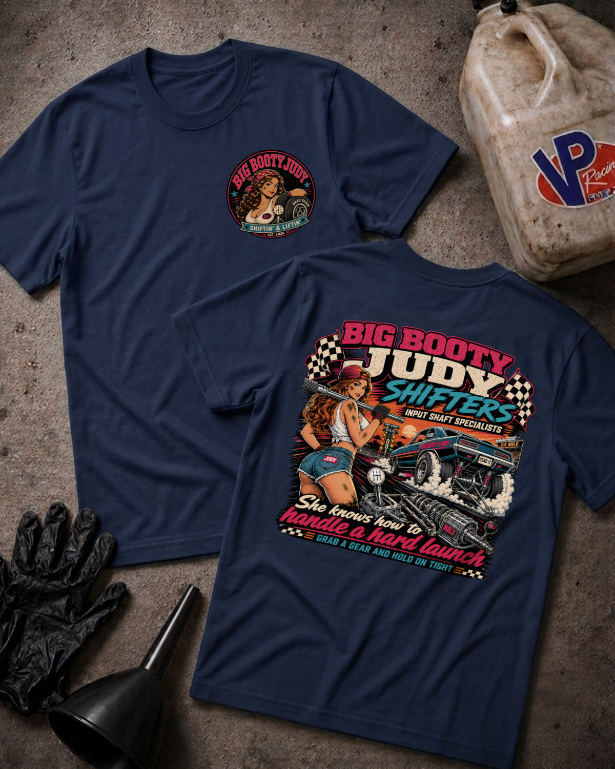 Two navy blue graphic T-shirts laid flat on a textured concrete-like surface, with racing and pin-up art graphics. One shirt shows a small circular chest print, and the other shows a large back graphic with the text 'BIG BOOTY JUDY SHIFTER' and a race car illustration.