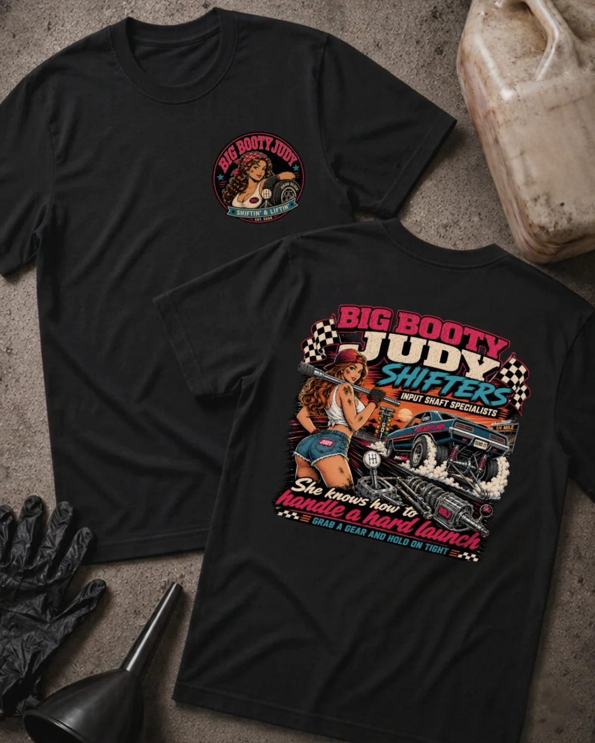 Two black  graphic T-shirts laid flat on a textured gray surface, with racing-themed artwork and text including 'BIG BOOTY JUDY SHIFTERS' and 'She knows how to handle a hard launch.' Props around them include a plastic gas can, black gloves, a funnel, and a worn bucket.