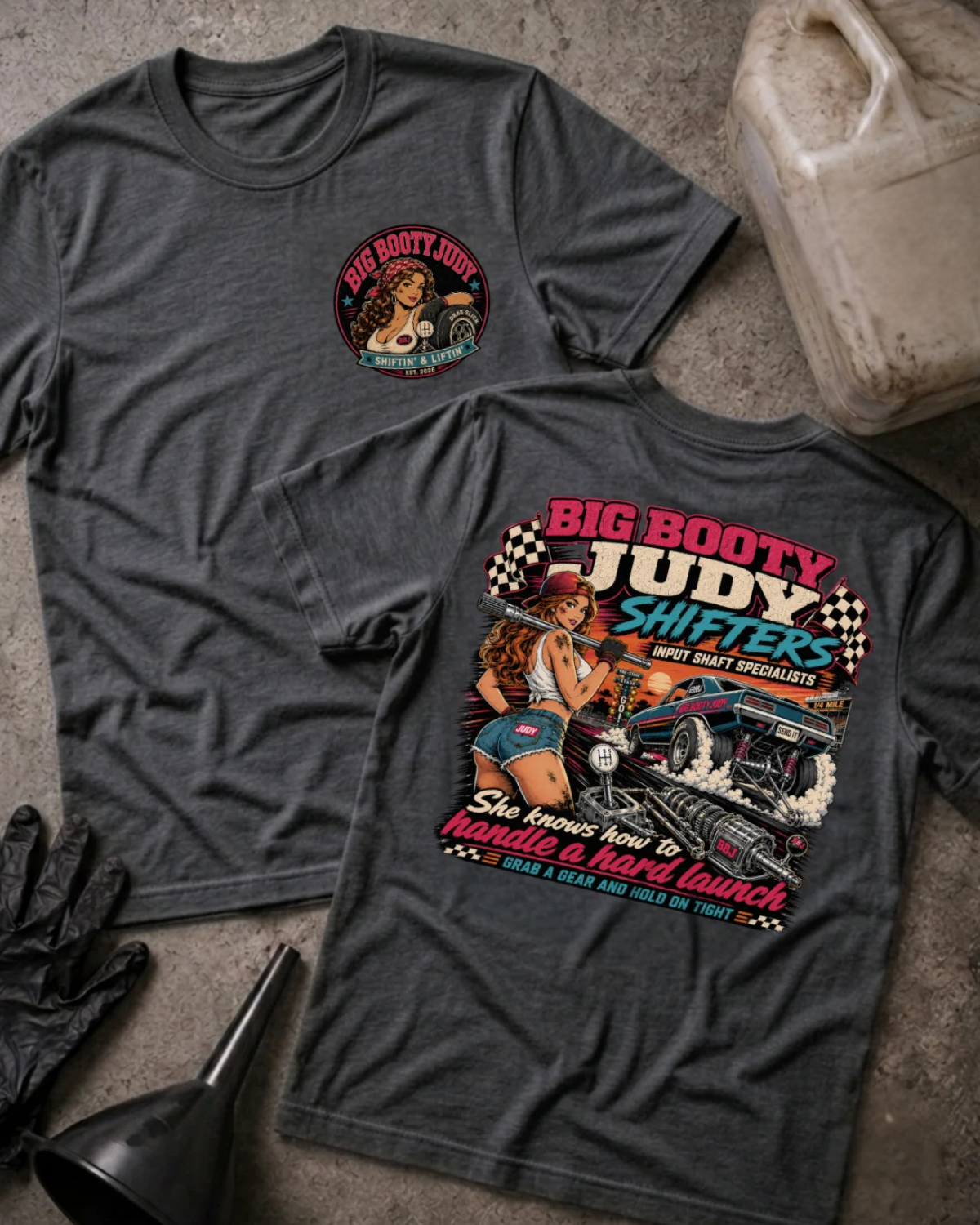 Two dark grey heather  graphic T-shirts laid flat on a textured gray surface, with racing-themed artwork and text including 'BIG BOOTY JUDY SHIFTERS' and 'She knows how to handle a hard launch.' Props around them include a plastic gas can, black gloves, a funnel, and a worn bucket.