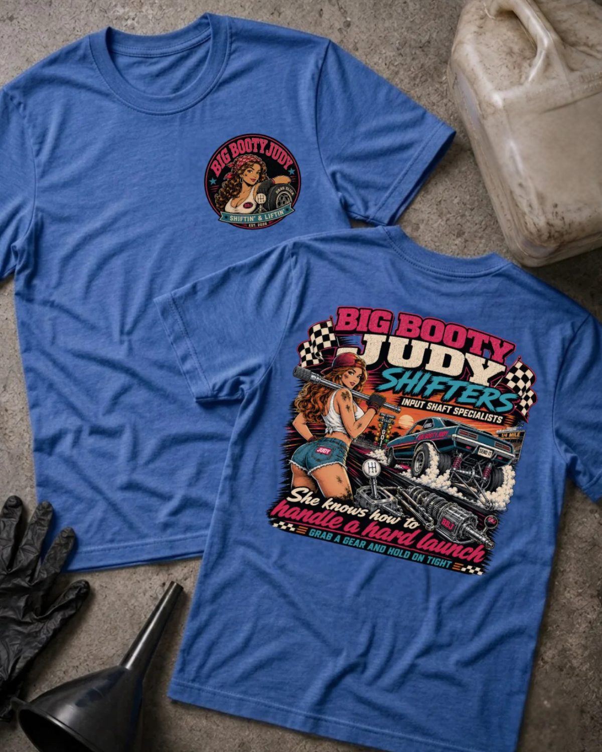 Two blue graphic T-shirts laid flat on a concrete surface, with a beige plastic jug, black gloves, and a black funnel as styling props. The front shirt shows a small circular chest graphic, and the back shirt shows a large colorful graphic with the text 'BIG BOOTY JUDY SHIFTERS' and an illustrated woman beside a race car.