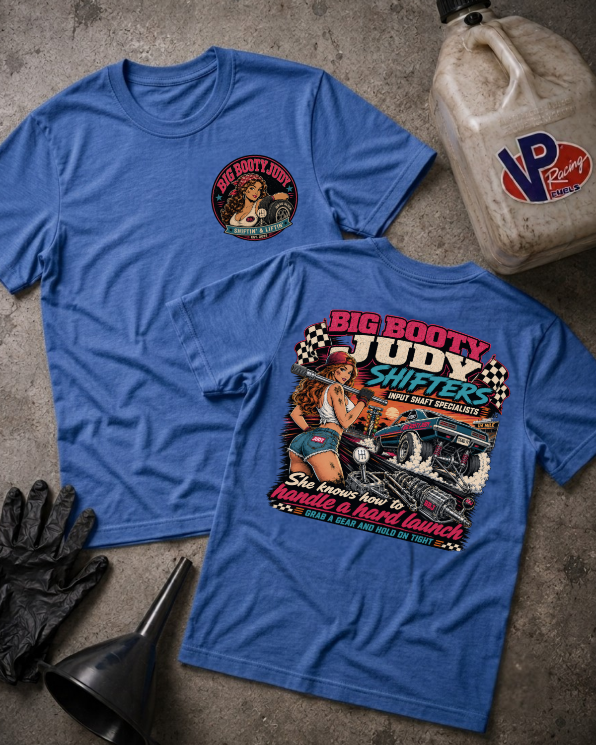 Two blue graphic T-shirts laid flat on a textured gray surface, with a vintage oil jug, black gloves, and a funnel around them. The front shirt has a small circular chest graphic, and the back shirt has a large retro-style racing pin-up graphic reading 'Big Booty Judy Shifters' with a driveshaft and car.