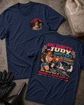 Two navy graphic T-shirts laid flat on a textured gray surface, with racing-themed artwork and text including 'BIG BOOTY JUDY SHIFTERS' and 'She knows how to handle a hard launch.' Props around them include a plastic gas can, black gloves, a funnel, and a worn bucket.