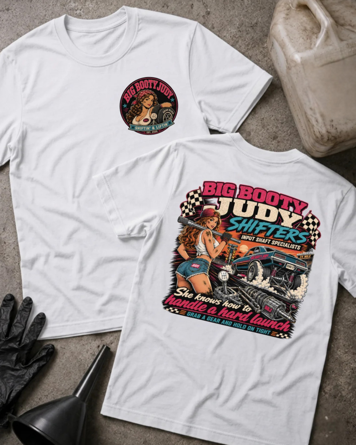 Two white  graphic T-shirts laid flat on a textured gray surface, with racing-themed artwork and text including 'BIG BOOTY JUDY SHIFTERS' and 'She knows how to handle a hard launch.' Props around them include a plastic gas can, black gloves, a funnel, and a worn bucket.