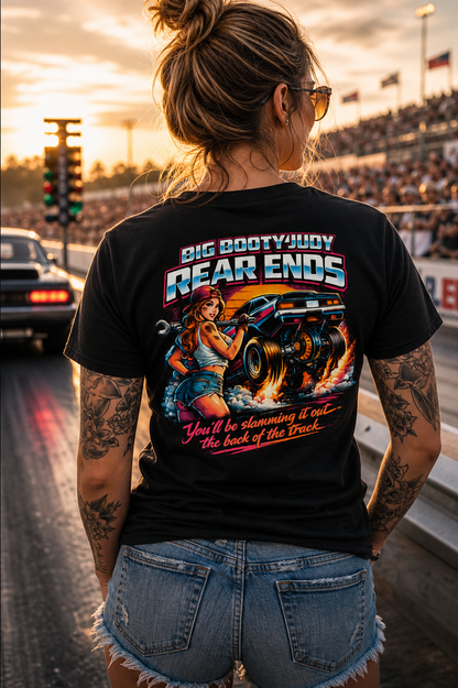Woman wearing a black Big Booty Judy Rear Ends t-shirt with a graphic design and cap, standing on a racetrack at sunset.