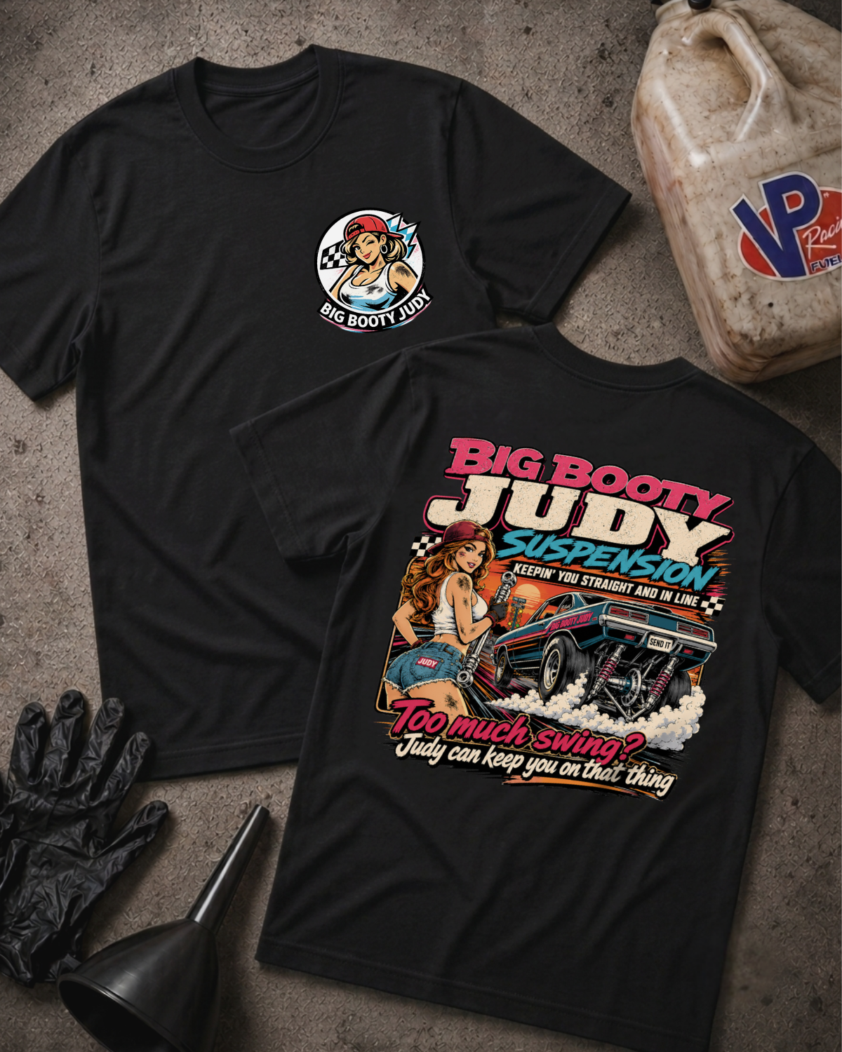 Two black graphic T-shirts laid flat on a textured gray surface, with automotive-themed printed artwork and text. One shirt shows a small circular chest graphic with a pin-up style illustration and the words 'BIG BOOTY JUDY'; the other shows a large back graphic with the same text in pink, white, and blue, plus an illustrated car and woman.
