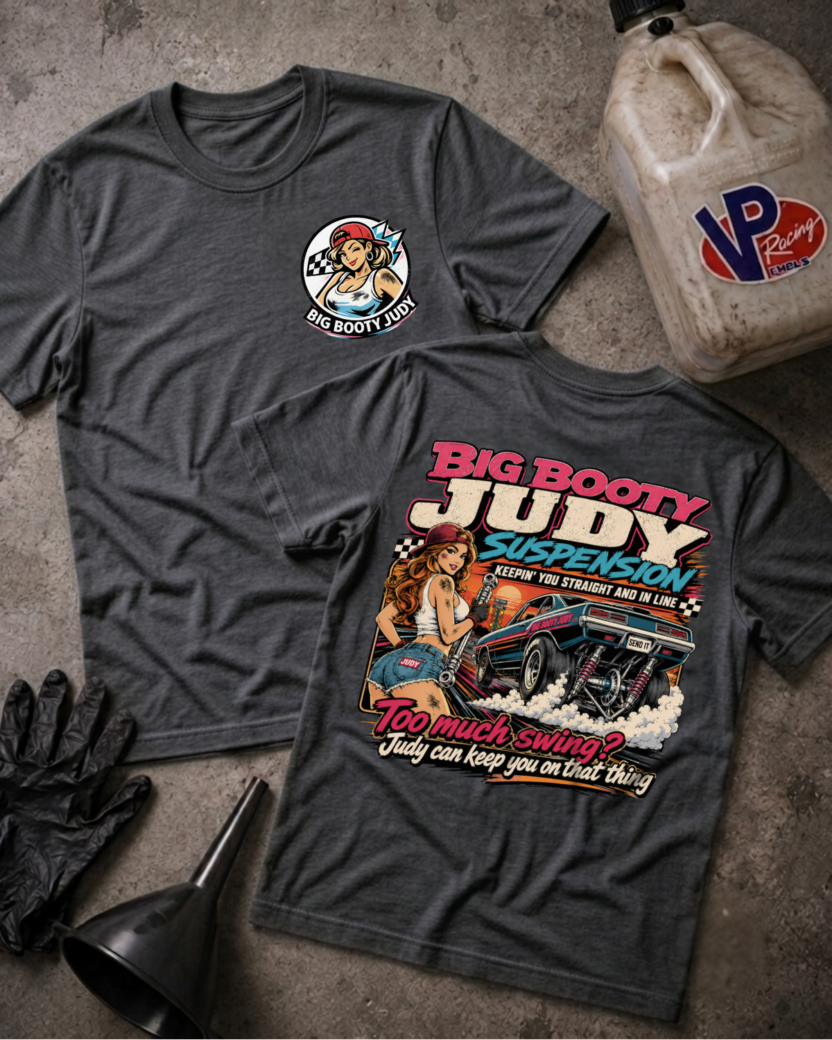 Two dark heather gray graphic T-shirts laid flat on a textured concrete surface, with a black glove, funnel, and a VP Racing fuel jug as styling props. The front shirt shows a small circular chest graphic, and the back shirt shows a large colorful graphic with the text 'BIG BOOTY JUDY SUSPENSION' and 'TOO MUCH SWING? JUDY CAN KEEP YOU ON THAT THING'
