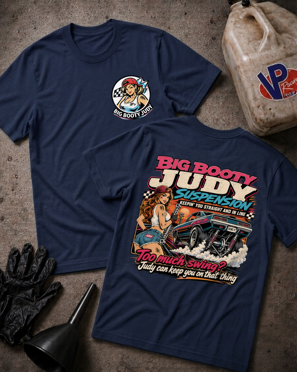 Two navy blue graphic T-shirts laid flat on a textured gray surface, with automotive-themed printed artwork and text including 'Big Booty Judy Suspenson' and 'Too much swing? Judy can keep you on that thing.' A plastic jug, black gloves, and funnel are placed around them as styled props.