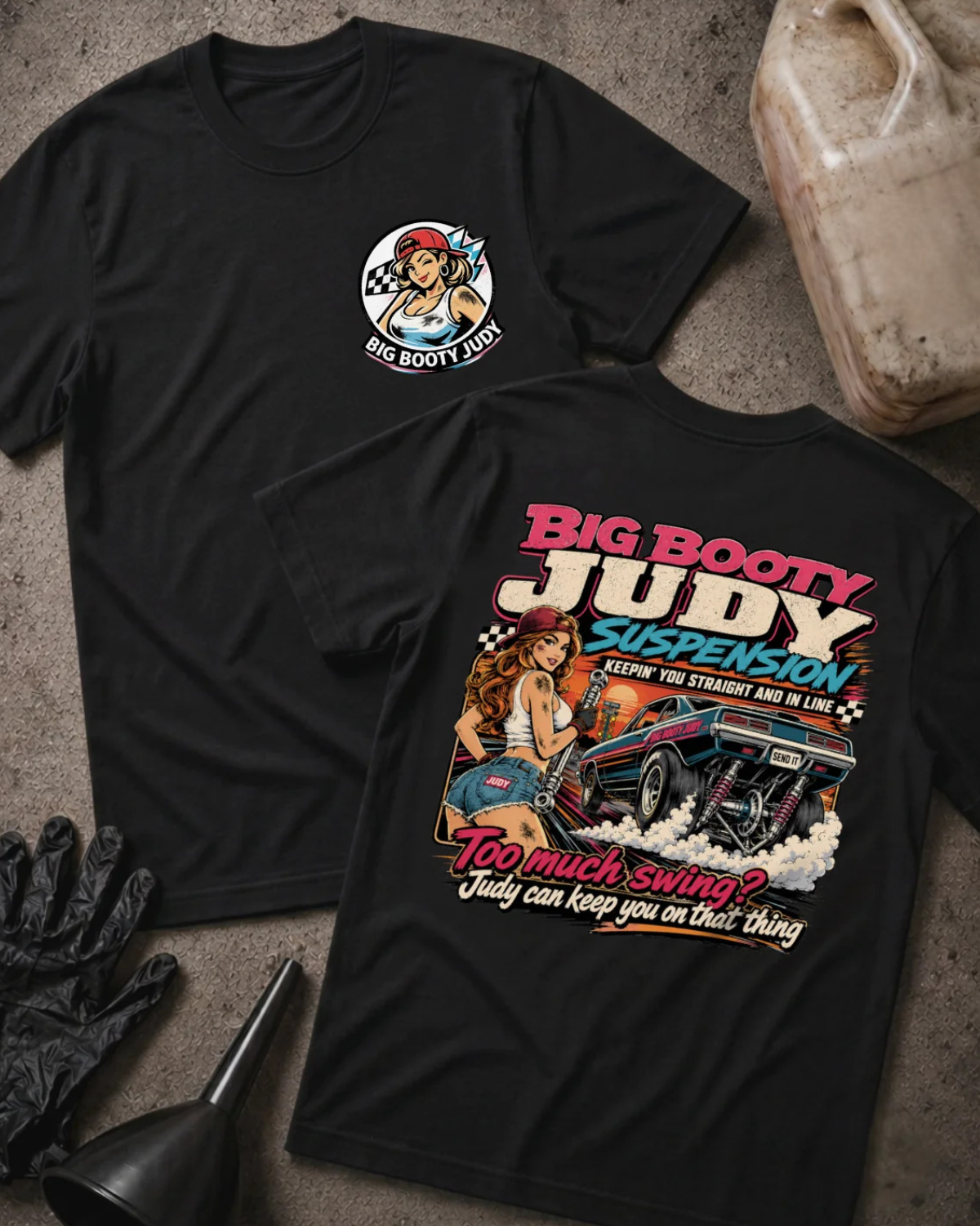 Two black graphic T-shirts laid flat on a textured concrete surface, with a black glove, funnel, and a VP Racing fuel jug as styling props. The front shirt shows a small circular chest graphic, and the back shirt shows a large colorful graphic with the text 'BIG BOOTY JUDY SUSPENSION' and 'TOO MUCH SWING? JUDY CAN KEEP YOU ON THAT THING'