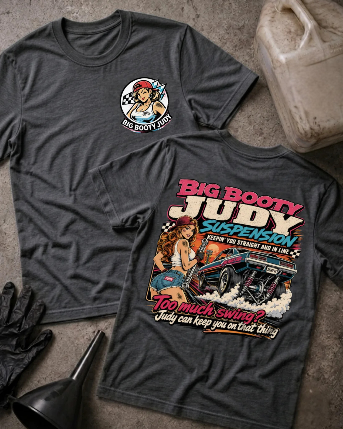 Two dark grey heather graphic T-shirts laid flat on a textured concrete surface, with a black glove, funnel, and a VP Racing fuel jug as styling props. The front shirt shows a small circular chest graphic, and the back shirt shows a large colorful graphic with the text 'BIG BOOTY JUDY SUSPENSION' and 'TOO MUCH SWING? JUDY CAN KEEP YOU ON THAT THING'