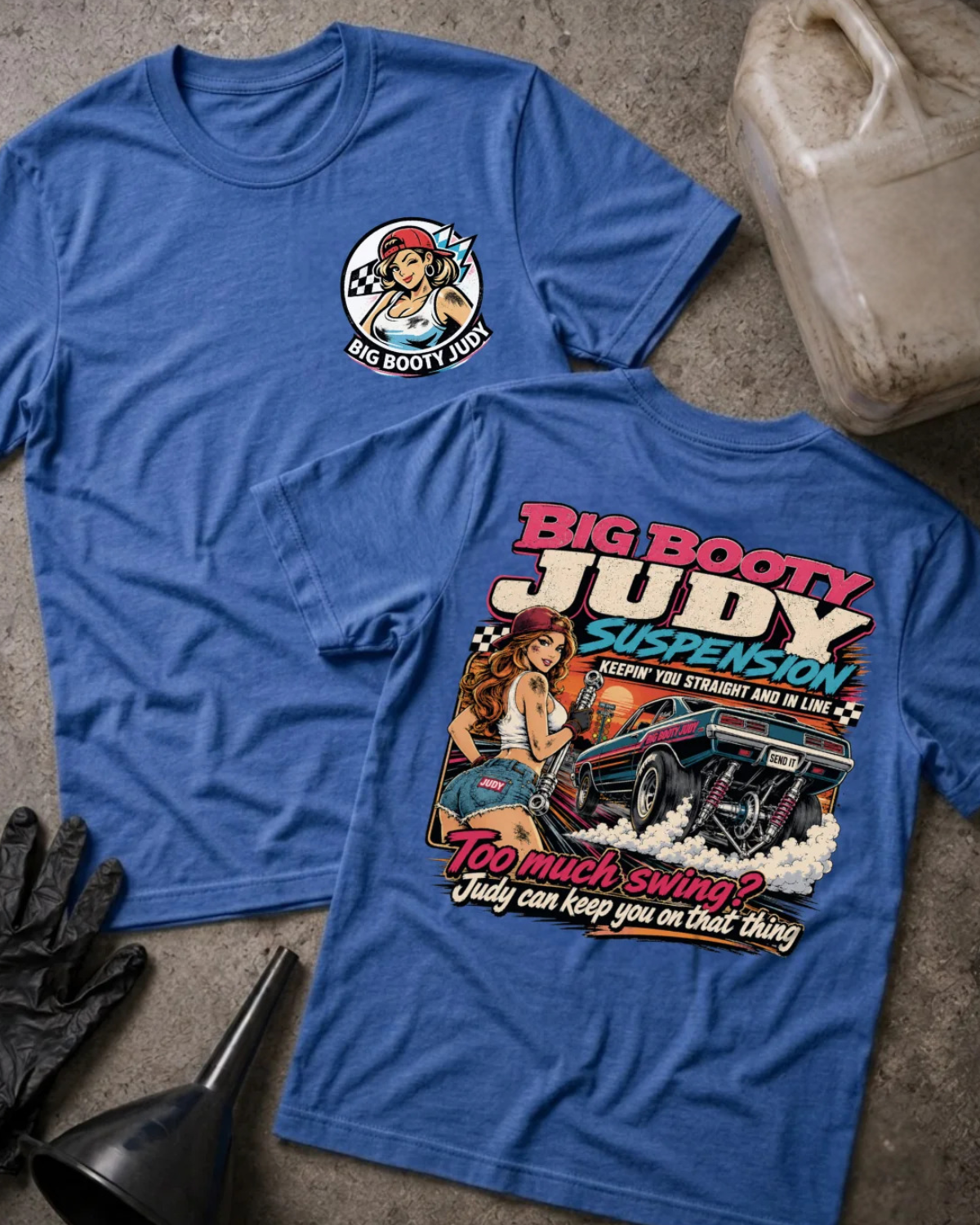 Two heather true royal graphic T-shirts laid flat on a textured concrete surface, with a black glove, funnel, and a VP Racing fuel jug as styling props. The front shirt shows a small circular chest graphic, and the back shirt shows a large colorful graphic with the text 'BIG BOOTY JUDY SUSPENSION' and 'TOO MUCH SWING? JUDY CAN KEEP YOU ON THAT THING'