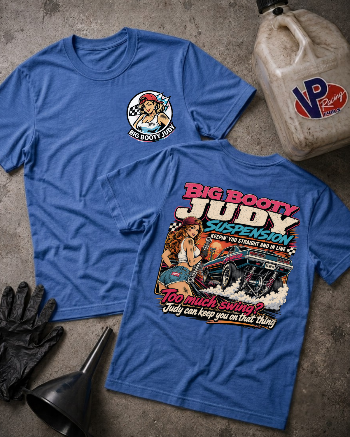 Two blue graphic T-shirts laid flat on a concrete floor, surrounded by styling props including a gas can labeled VP Racing, black gloves, and a funnel. The front shirt shows a large retro-style graphic with the text 'Big Booty Judy Suspension' and a car illustration.