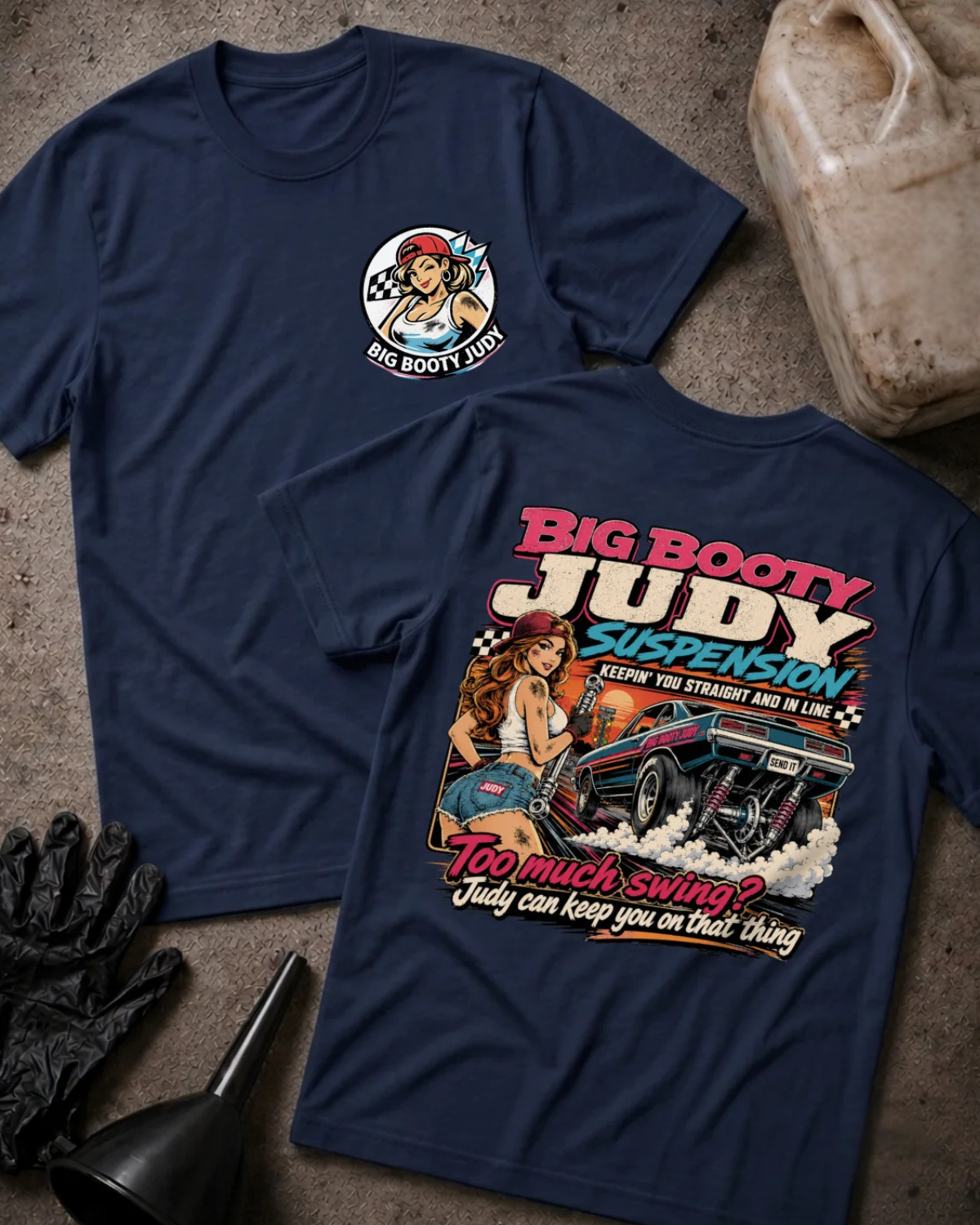 Two navy graphic T-shirts laid flat on a textured concrete surface, with a black glove, funnel, and a VP Racing fuel jug as styling props. The front shirt shows a small circular chest graphic, and the back shirt shows a large colorful graphic with the text 'BIG BOOTY JUDY SUSPENSION' and 'TOO MUCH SWING? JUDY CAN KEEP YOU ON THAT THING'