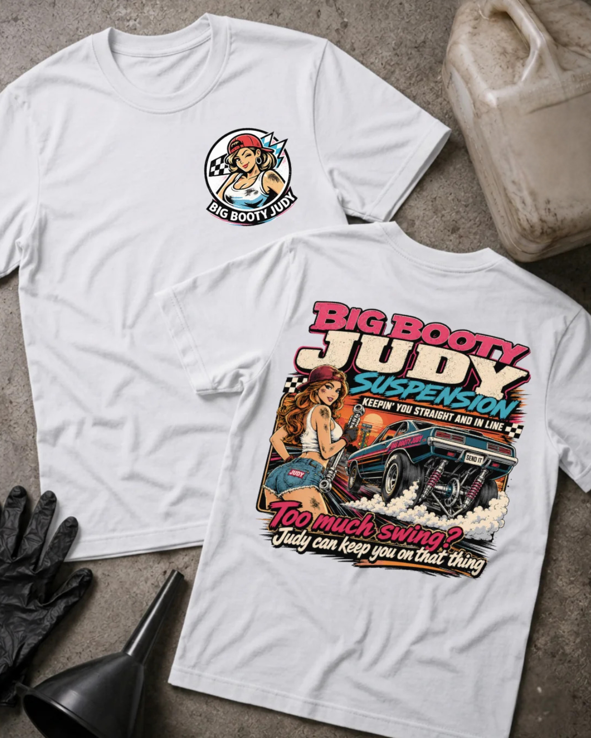 Two white graphic T-shirts laid flat on a textured concrete surface, with a black glove, funnel, and a VP Racing fuel jug as styling props. The front shirt shows a small circular chest graphic, and the back shirt shows a large colorful graphic with the text 'BIG BOOTY JUDY SUSPENSION' and 'TOO MUCH SWING? JUDY CAN KEEP YOU ON THAT THING'