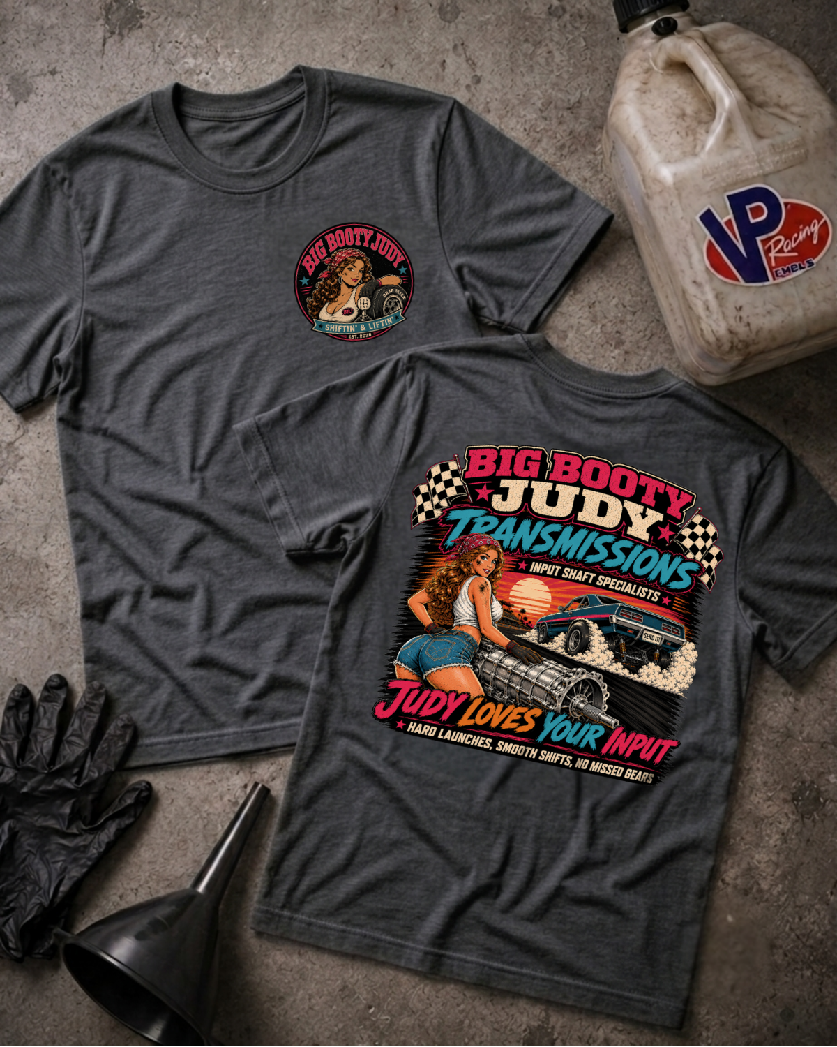 Two dark heather gray graphic T-shirts laid flat on a textured concrete surface, with a black glove, funnel, and a VP Racing fuel jug as styling props. The front shirt shows a small circular chest graphic, and the back shirt shows a large colorful graphic with the text 'BIG BOOTY JUDY TRANSMISSIONS' and 'JUDY LOVES YOUR INPUT.