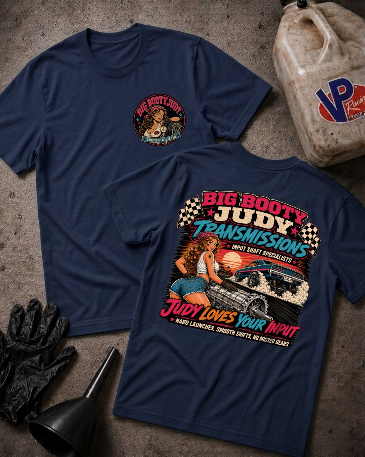Two navy blue graphic T-shirts laid flat on a textured gray surface, with automotive-themed printed artwork and text including 'Big Booty Judy Transmissions' and 'Judy Loves Your Input.' A plastic jug, black gloves, and funnel are placed around them as styled props.