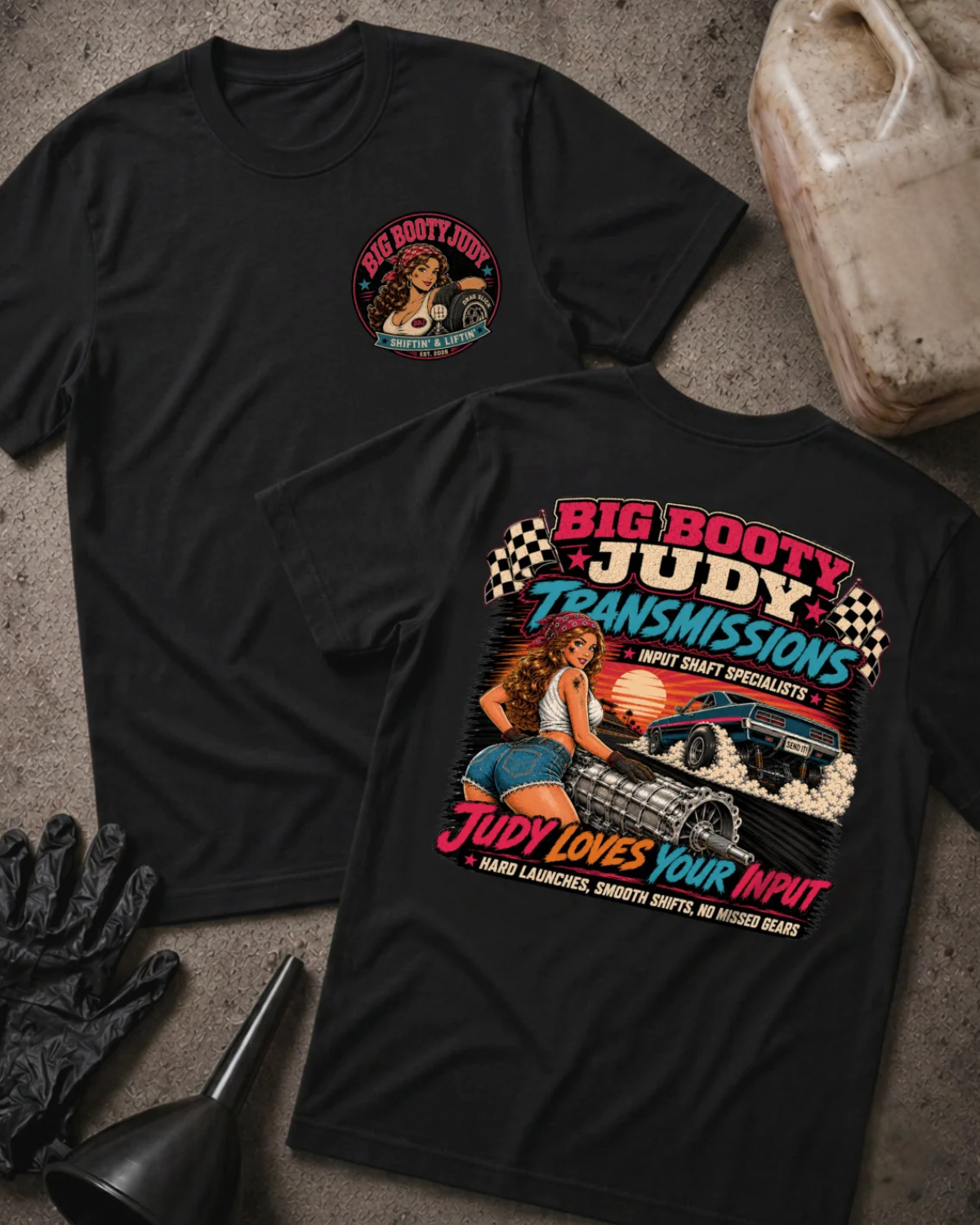 Two black graphic T-shirts laid flat on a textured gray surface, with automotive-themed printed artwork and 'Big Booty Judy Transmissions Judy Loves Your Input' text. A plastic fuel can, black gloves, and a funnel are placed around them as styled props
