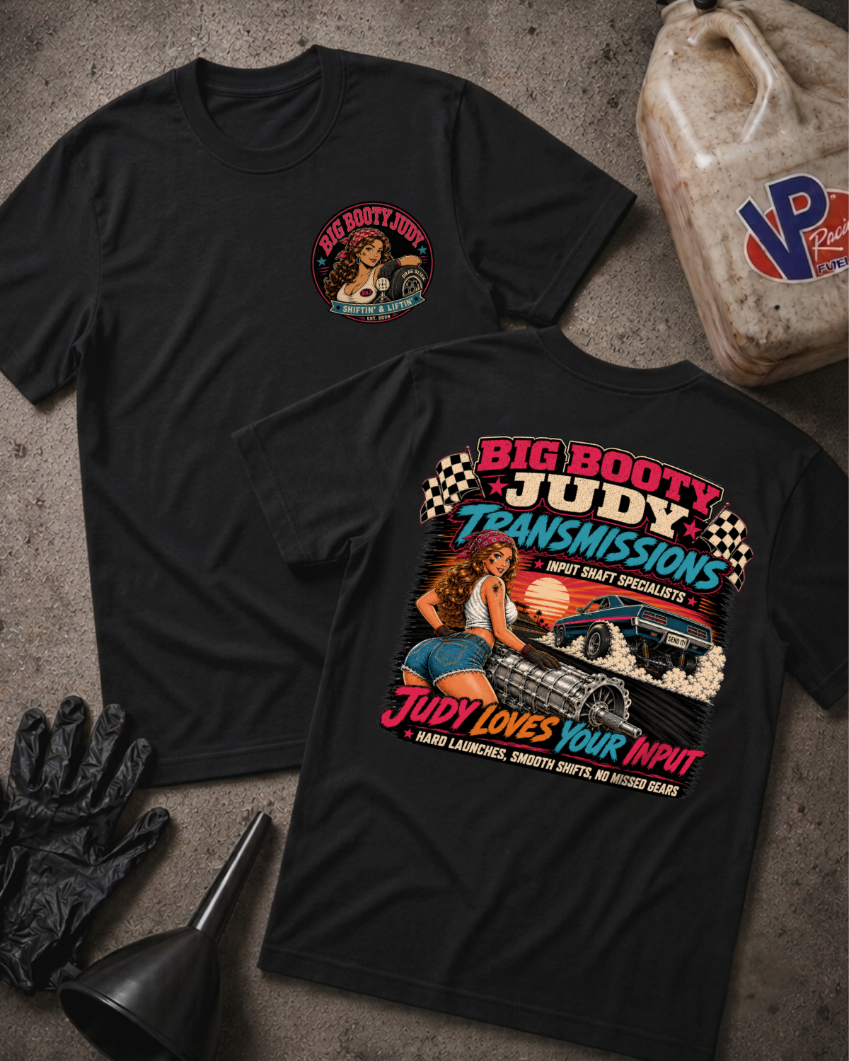 Two black graphic T-shirts laid flat on a textured gray surface, with automotive-themed printed artwork and 'Big Booty Judy Transmissions Judy Loves Your Input' text. A plastic fuel can, black gloves, and a funnel are placed around them as styled props.