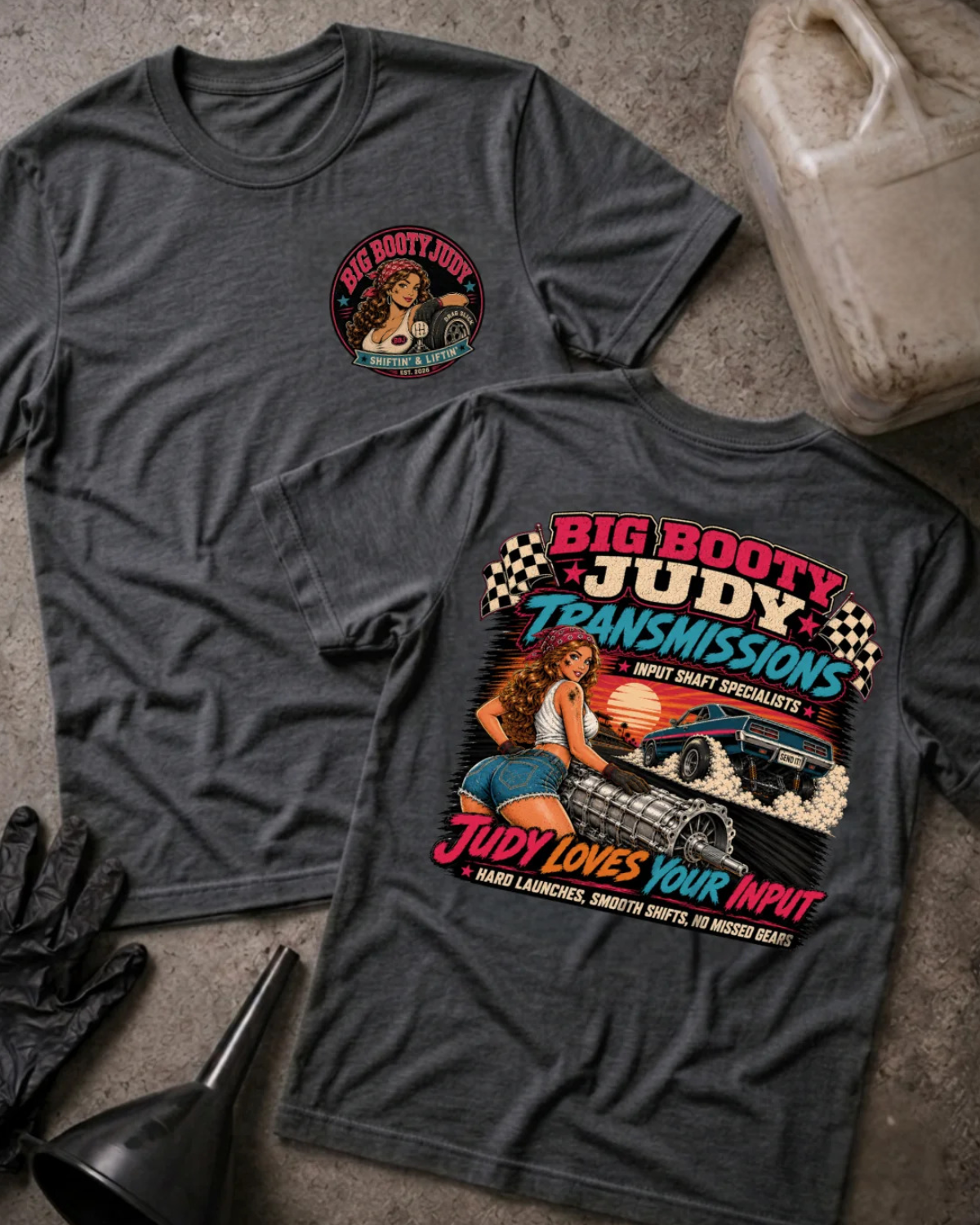 Two dark heather gray graphic T-shirts laid flat on a concrete-like surface with automotive props. One shirt shows a small circular chest graphic, and the other shows a large front graphic with the text 'Big Booty Judy Transmissions' and an illustrated pin-up car scene.