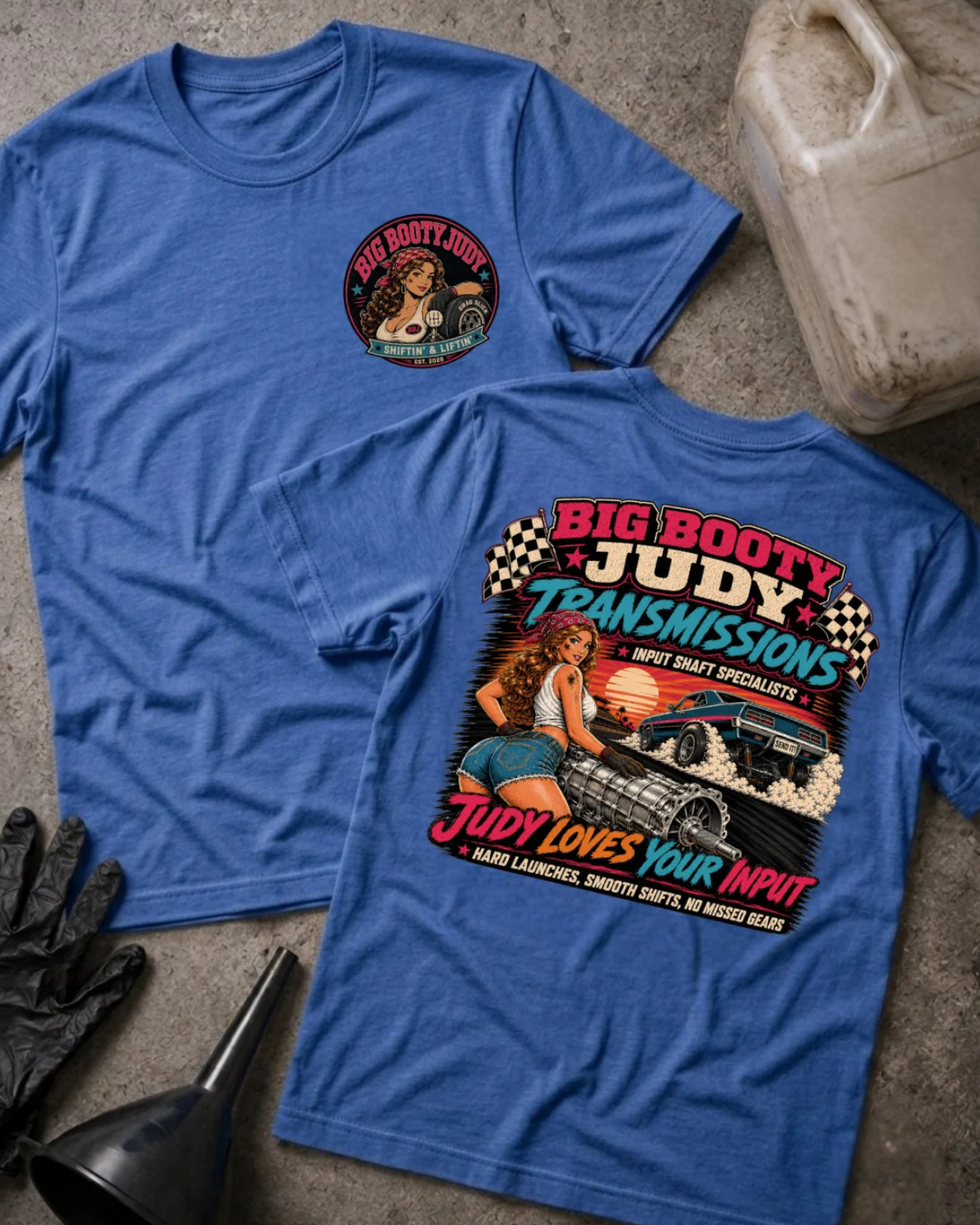 Two blue graphic T-shirts laid flat on a gray concrete-like surface, showing the front and back designs. The front shirt has a small circular chest graphic, and the back shirt has a large colorful graphic with the text 'BIG BOOTY JUDY TRANSMISSIONS' and 'JUDY LOVES YOUR INPUT.