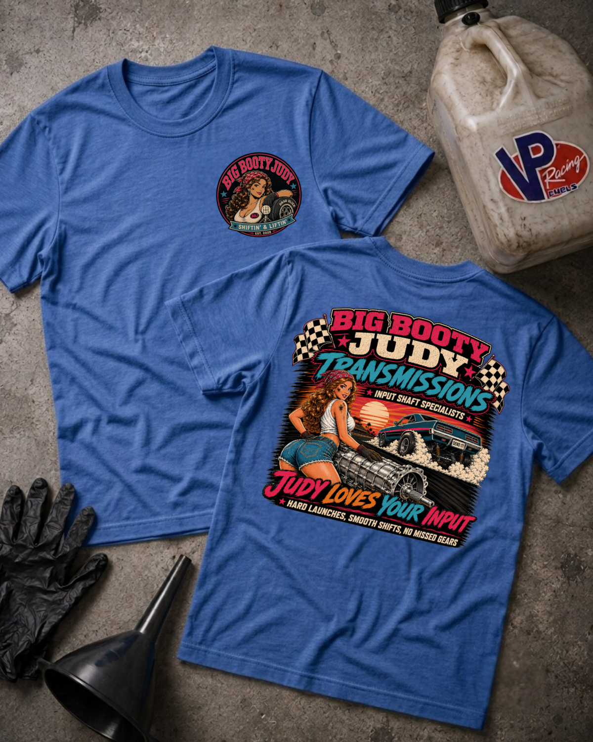 Two blue graphic T-shirts laid flat on a concrete surface, one showing the front with a small circular chest print and the other showing the back with a large retro-style automotive graphic. A plastic fuel jug, black gloves, and a funnel are placed around them as styled props.