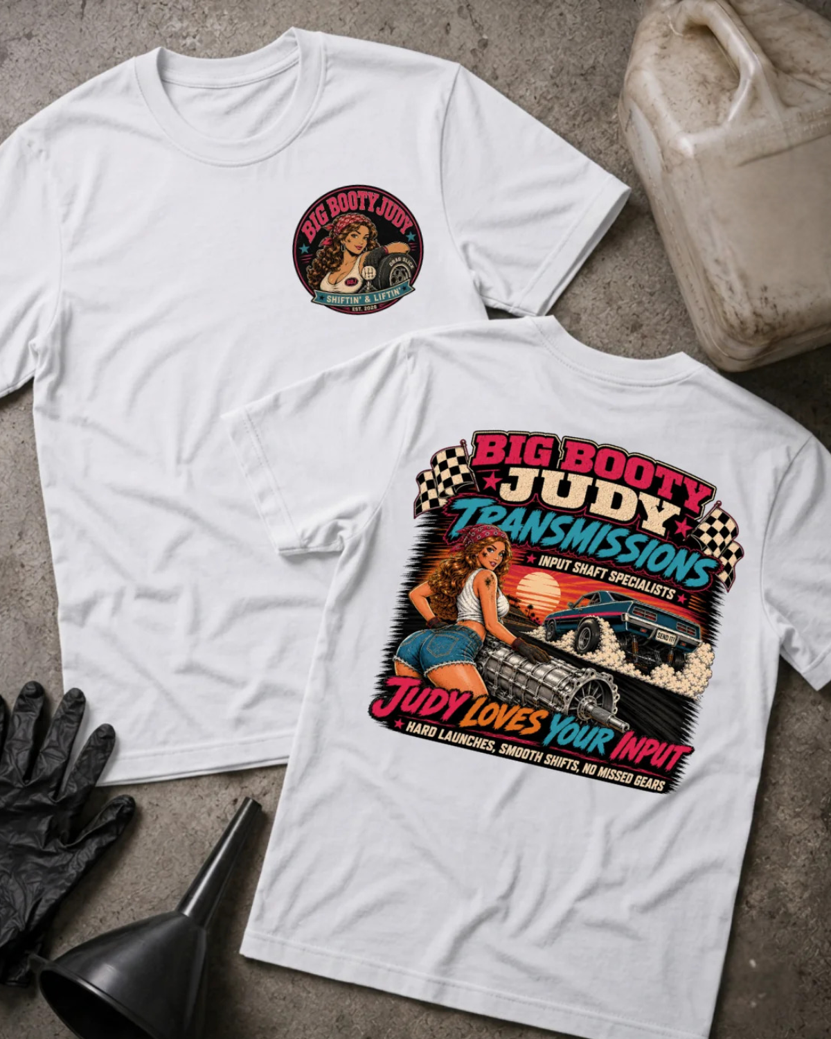 Two white graphic T-shirts laid flat on a gray concrete surface with automotive props. The front shirt shows a circular chest graphic, and the back shirt shows a large colorful illustration with the text 'BIG BOOTY JUDY TRANSMISSIONS' and 'JUDY LOVES YOUR INPUT.