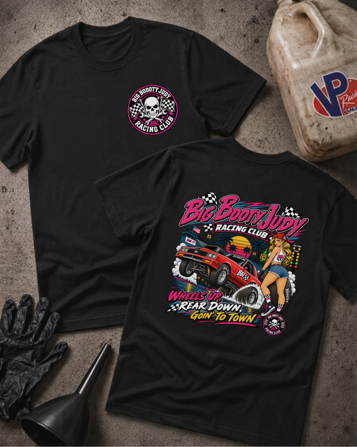 Two black graphic T-shirts laid flat on a textured gray surface, with a black oil funnel, black gloves, and a worn plastic jerrycan nearby. The front shirt shows a small circular chest graphic with a skull and the words 'Big Booty Racing Club.