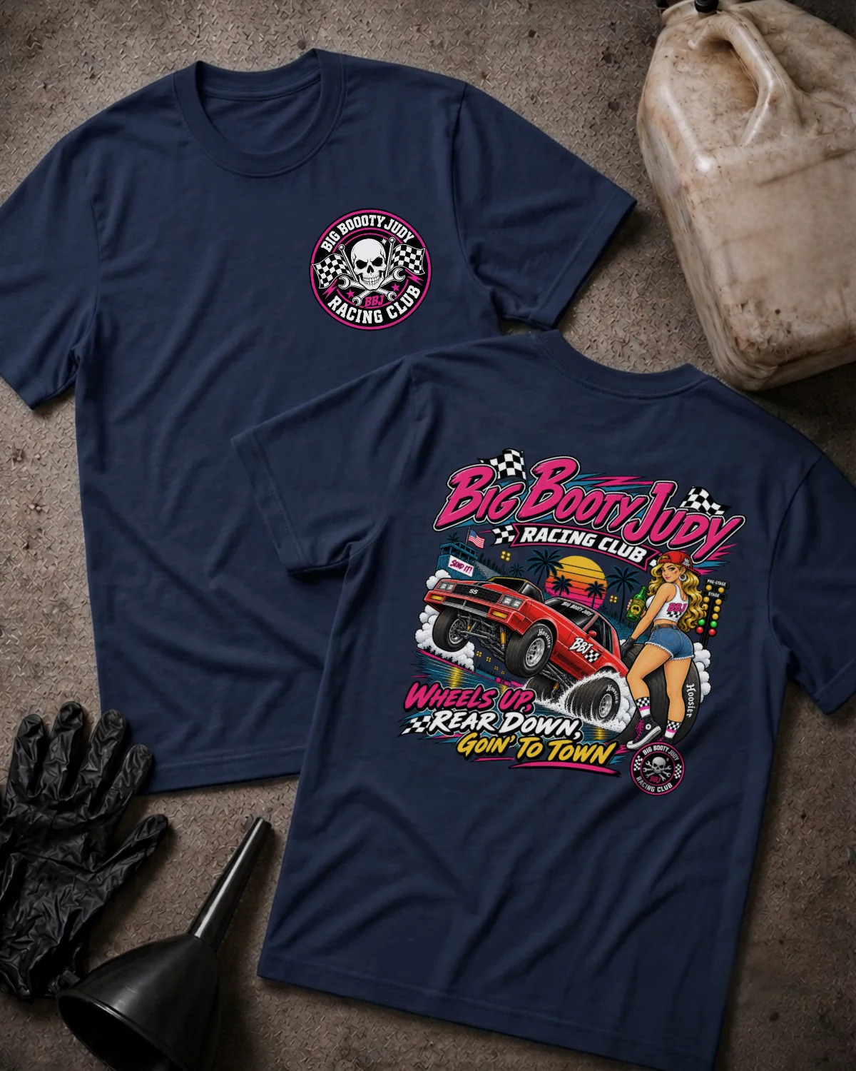 Two navy graphic t-shirts displayed on a textured gray surface with racing-themed props, including a jerrycan, gloves, and funnel. The front shirt features a circular 'Big Booty Judy Racing Club' logo on the left chest, while the back shirt displays a large, colorful racing graphic with the text 'Big Boot Judy Racing Club' and 'Wheels Up, Rear Down, Goin To Town'.