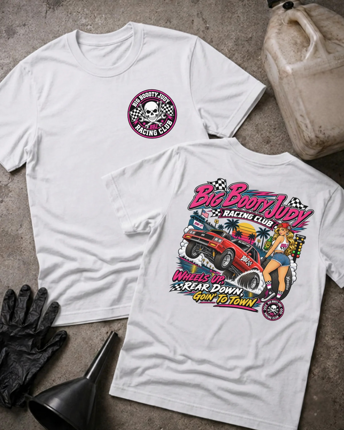 Two white graphic t-shirts displayed on a textured gray surface with racing-themed props, including a jerrycan, gloves, and funnel. The front shirt features a circular 'Big Booty Judy Racing Club' logo on the left chest, while the back shirt displays a large, colorful racing graphic with the text 'Big Boot Judy Racing Club' and 'Wheels Up, Rear Down, Goin To Town'.