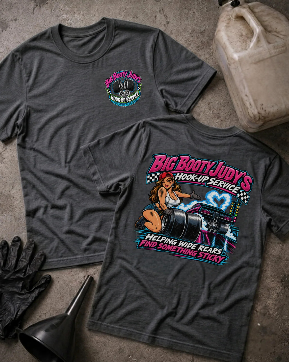 Two dark heather gray graphic T-shirts laid flat on a concrete surface, with a black funnel, black gloves, and a dirty plastic jug as styling props. The front shirt shows a small circular chest graphic, and the back shirt shows a large colorful retro-style graphic with the text 'Big Booty Judy's Hookup Service' and 'Helping Ride Rears and Something Sticky.