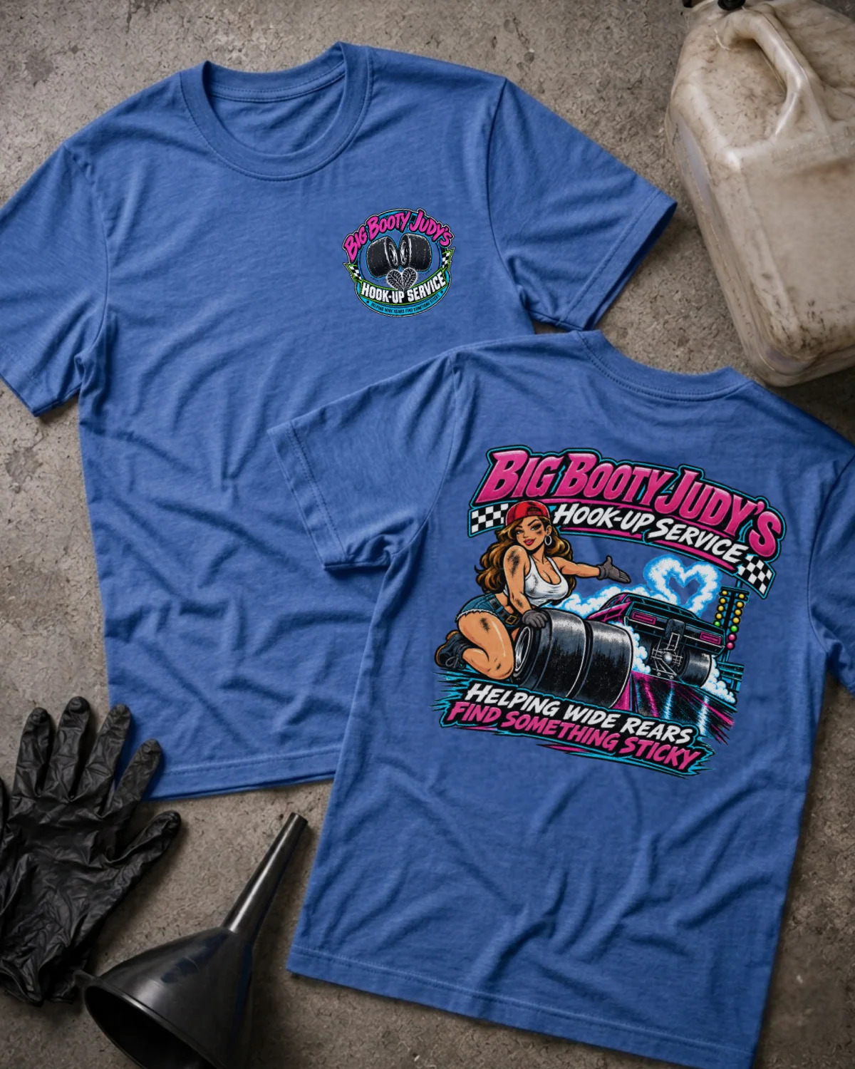 Two blue graphic T-shirts laid flat on a concrete surface with automotive props. The front shirt shows a small circular chest graphic, and the back shirt shows a large colorful graphic with the text 'Big Booty Judy's Hook-Up Service' and an illustrated woman beside a car.