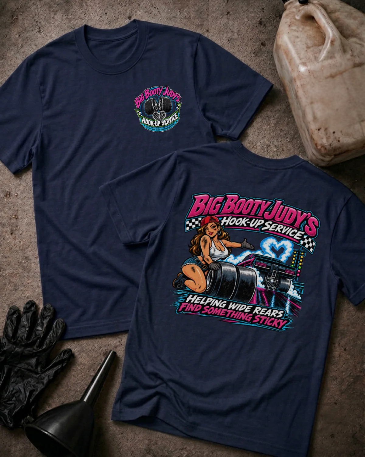 Two navy blue graphic T-shirts laid flat on a rustic concrete-like surface, with automotive-themed props including a dirty plastic jug, black gloves, and a funnel. The front shirt shows a small circular chest graphic, and the back shirt shows a large colorful retro-style graphic with the text 'Big Booty Judy's Hook-Up Service' and 'Helping Wide Rears And Something Sticky.
