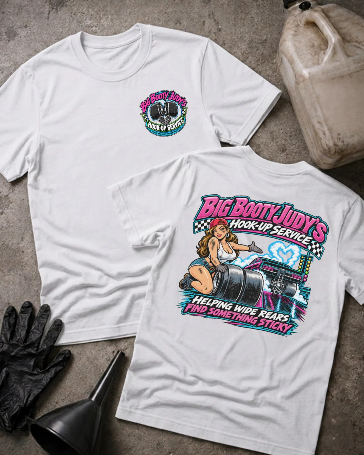 Two white graphic T-shirts laid flat on a concrete surface, showing the front and back designs. The front shirt has a small circular chest graphic, and the back shirt has a large colorful graphic with the text 'Big Boot Judy's Hook-Up Service' and an illustration.