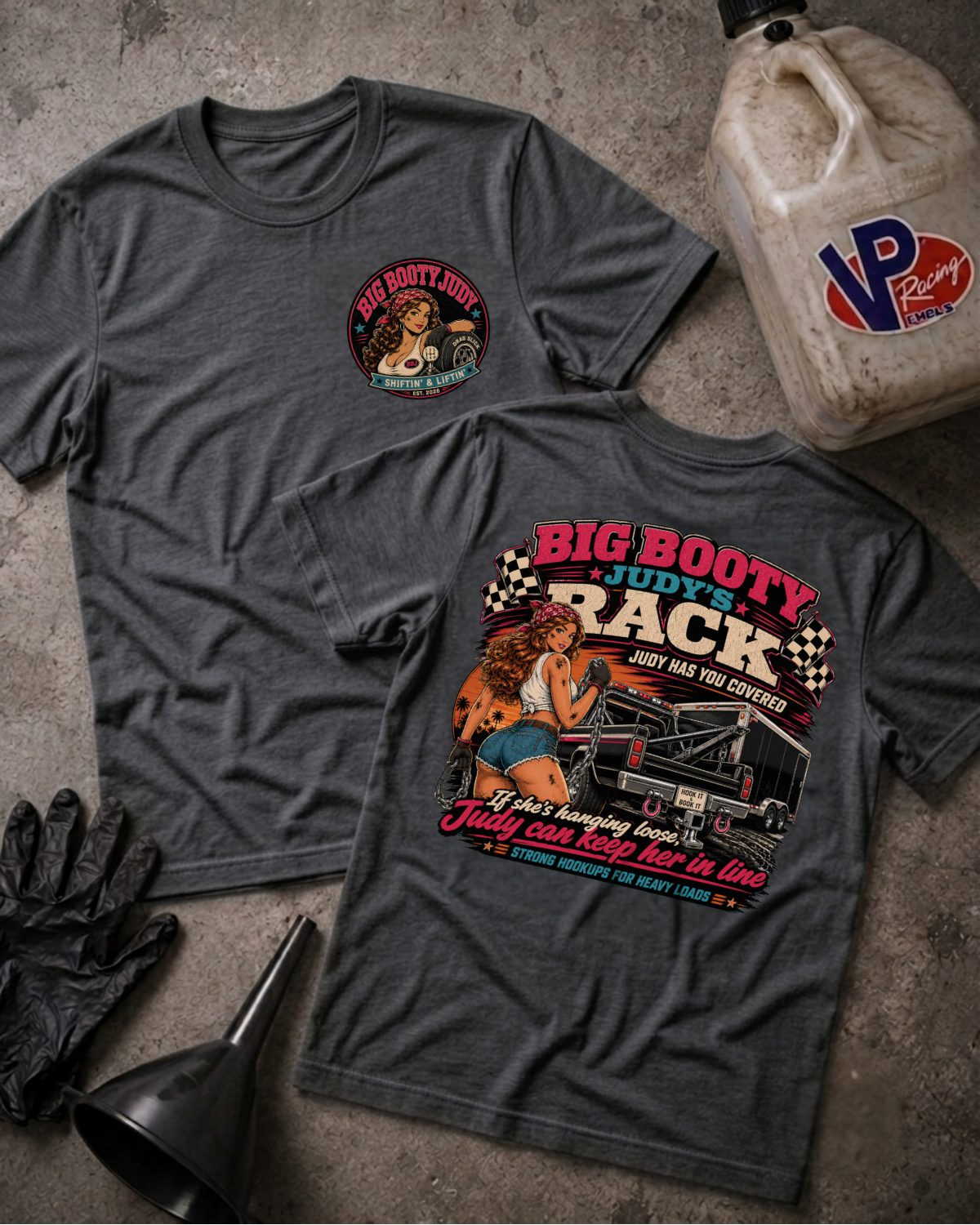 Two dark heather gray graphic t-shirts displayed on a textured concrete floor with garage-themed props, including a VP Racing jug, black gloves, and a funnel. One shirt shows a small circular graphic on the left chest, and the other shows a large back graphic featuring a woman driving a race car with the text 'BIG BOOTY JUDY'S RACK'.
