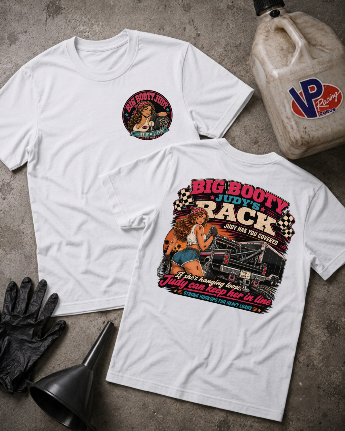 Two white graphic T-shirts laid flat on a gray concrete-style surface, with a black funnel, black gloves, and a plastic jug nearby. The front shirt shows a small circular chest graphic, and the back shirt shows a large colorful racing-themed print with the text 'BIG BOOTY JUDY'S RACK' and an illustrated woman beside a race car.