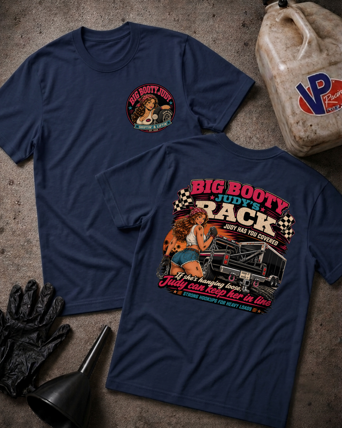 Two navy blue graphic T-shirts laid flat on a textured gray surface, with automotive-themed printed artwork and text including 'Big Booty Judy's Rack.' A plastic jug, black gloves, and funnel are placed around them as styled props.