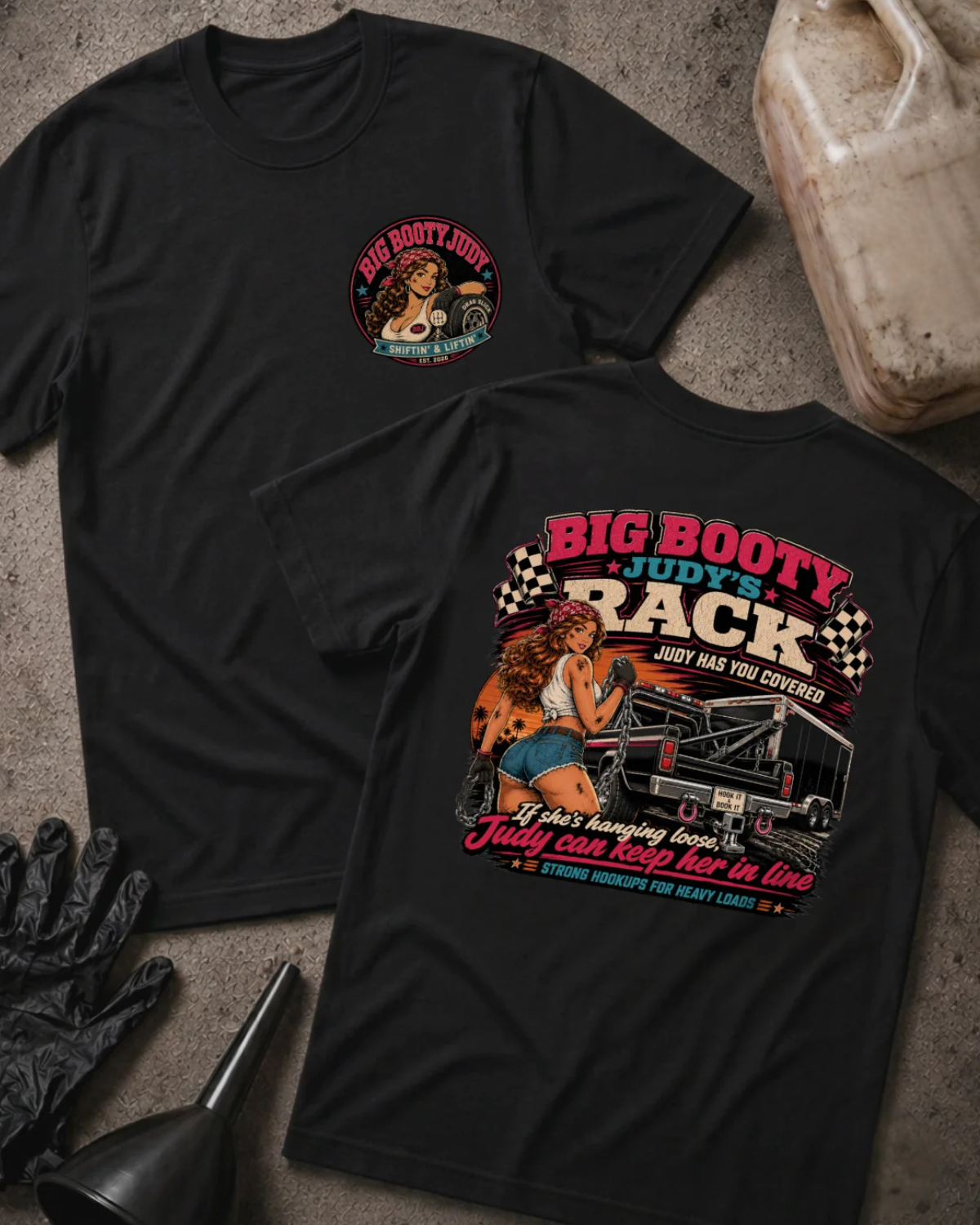 Two black graphic T-shirts laid flat on a gray concrete-style surface, with a black funnel, black gloves, and a plastic jug nearby. The front shirt shows a small circular chest graphic, and the back shirt shows a large colorful racing-themed print with the text 'BIG BOOTY JUDY'S RACK' and an illustrated woman beside a race car.