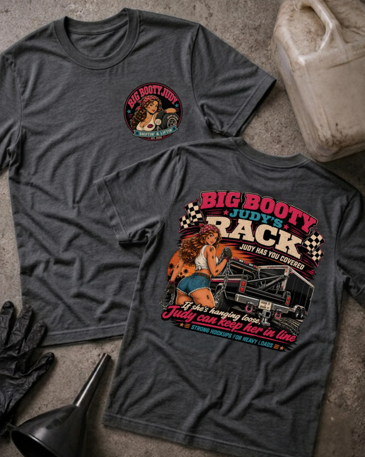Two dark heather gray graphic T-shirts laid flat on a concrete floor, showing both front and back designs. The front has a small circular chest graphic, and the back has a large colorful graphic with the text 'BIG BOOTY JUDY'S RACK' and an illustration.