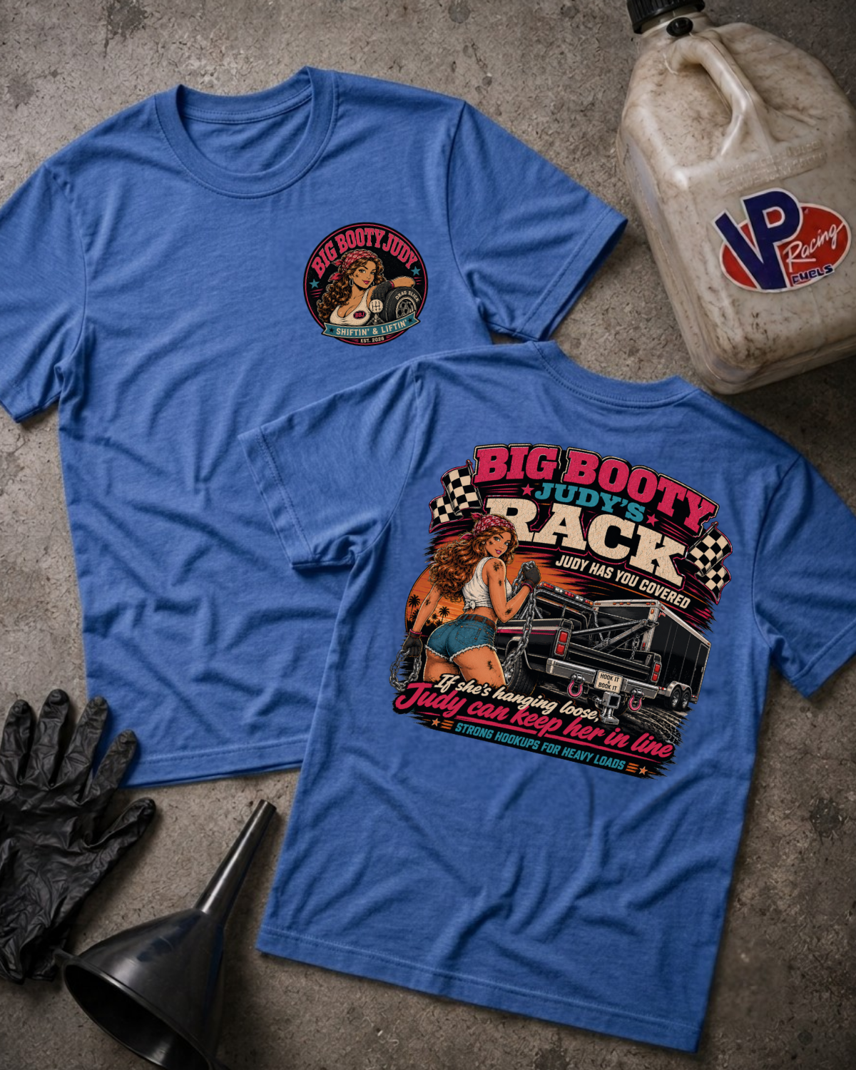 Two blue graphic T-shirts laid flat on a textured gray surface, with a vintage oil can, black gloves, and a black funnel around them. The front shirt has a small circular chest graphic, and the back shirt has a large retro-style racing graphic with the text 'BIG BOOTY JUDY'S RACK' and an illustrated woman beside a race car