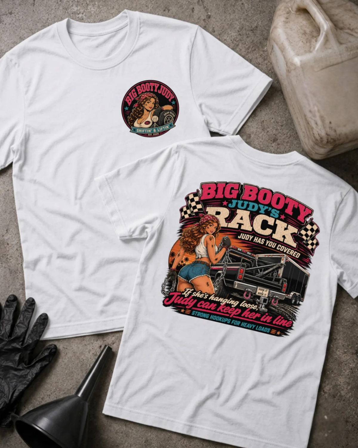Two white graphic T-shirts laid flat on a gray concrete surface, with a black funnel, black gloves, and a plastic jug as styling props. The front shirt shows a small circular chest graphic, and the back shirt shows a large retro-style graphic with the text 'BIG BOOTY JUDY'S RACK' and pin-up art.