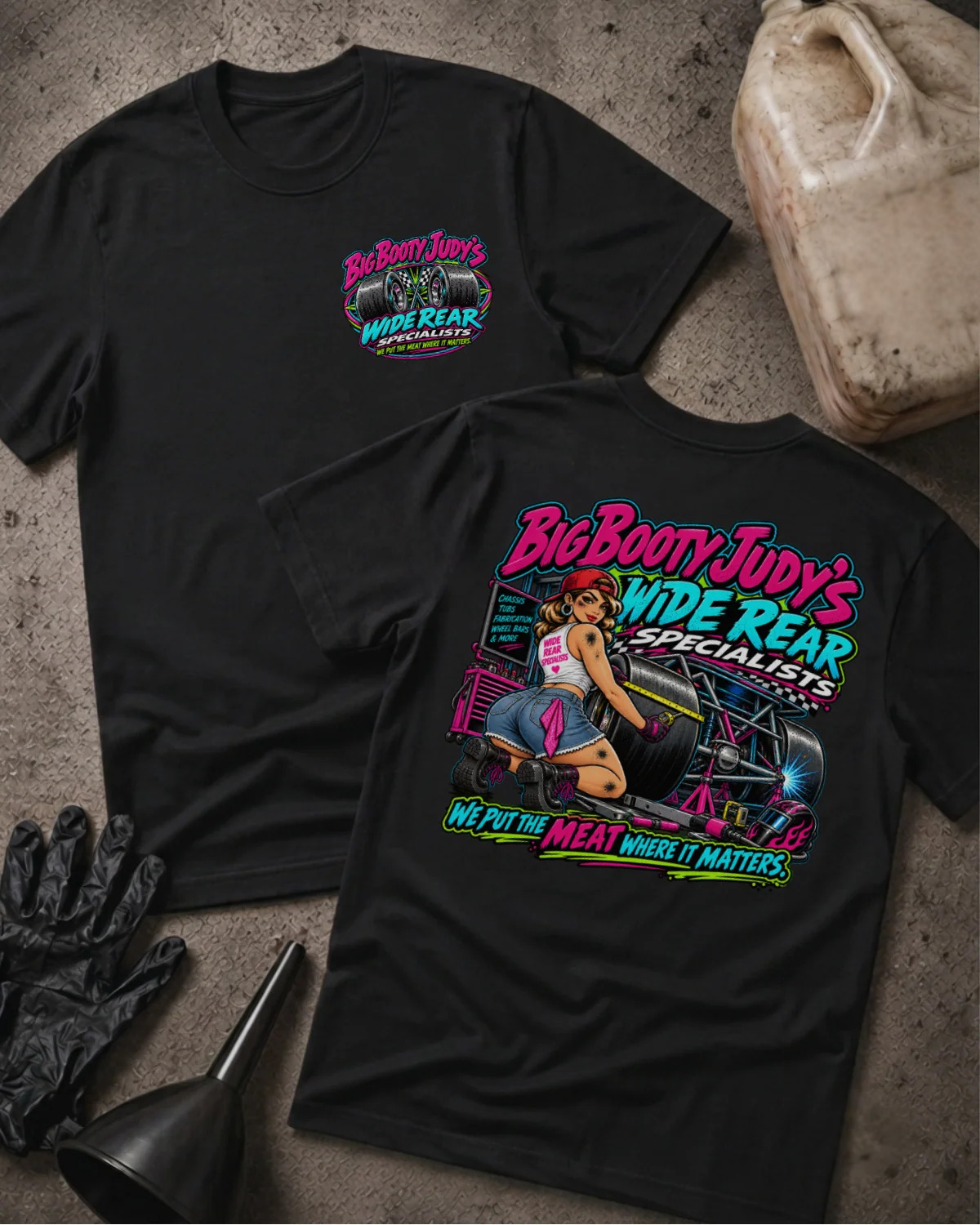 Two black graphic T-shirts laid flat on a textured concrete surface, showing the front and back designs. The shirts feature a colorful 'Big Boony Judy's Wide Rear Specialists' graphic with a pin-up illustration and neon-style text.