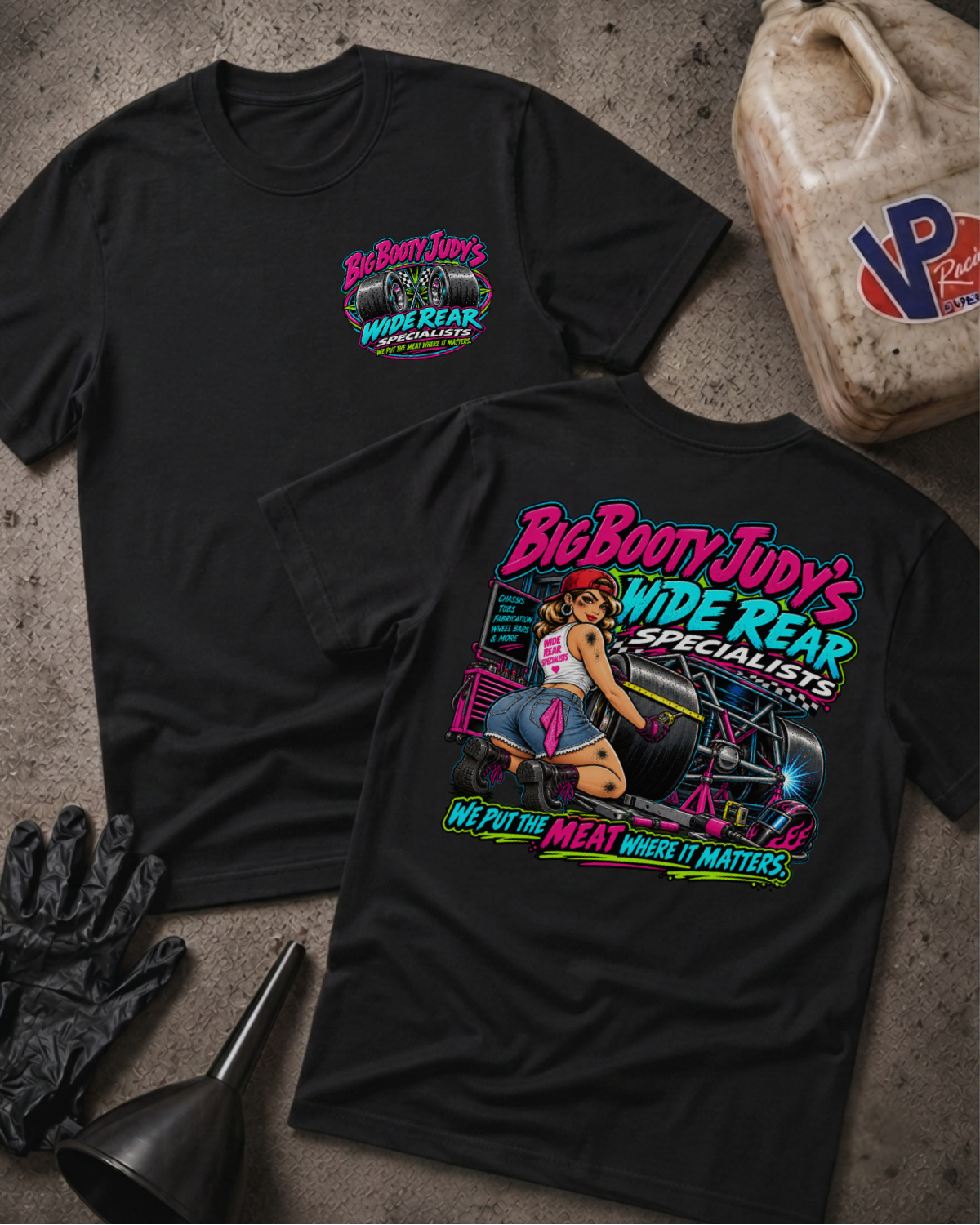 Two black graphic T-shirts laid flat on a textured concrete-like surface, with automotive props including a plastic jug, gloves, funnel, and brush around them. The front shirt shows a small multicolor chest logo reading Big Booty Judy's Wide Rear Specialists.