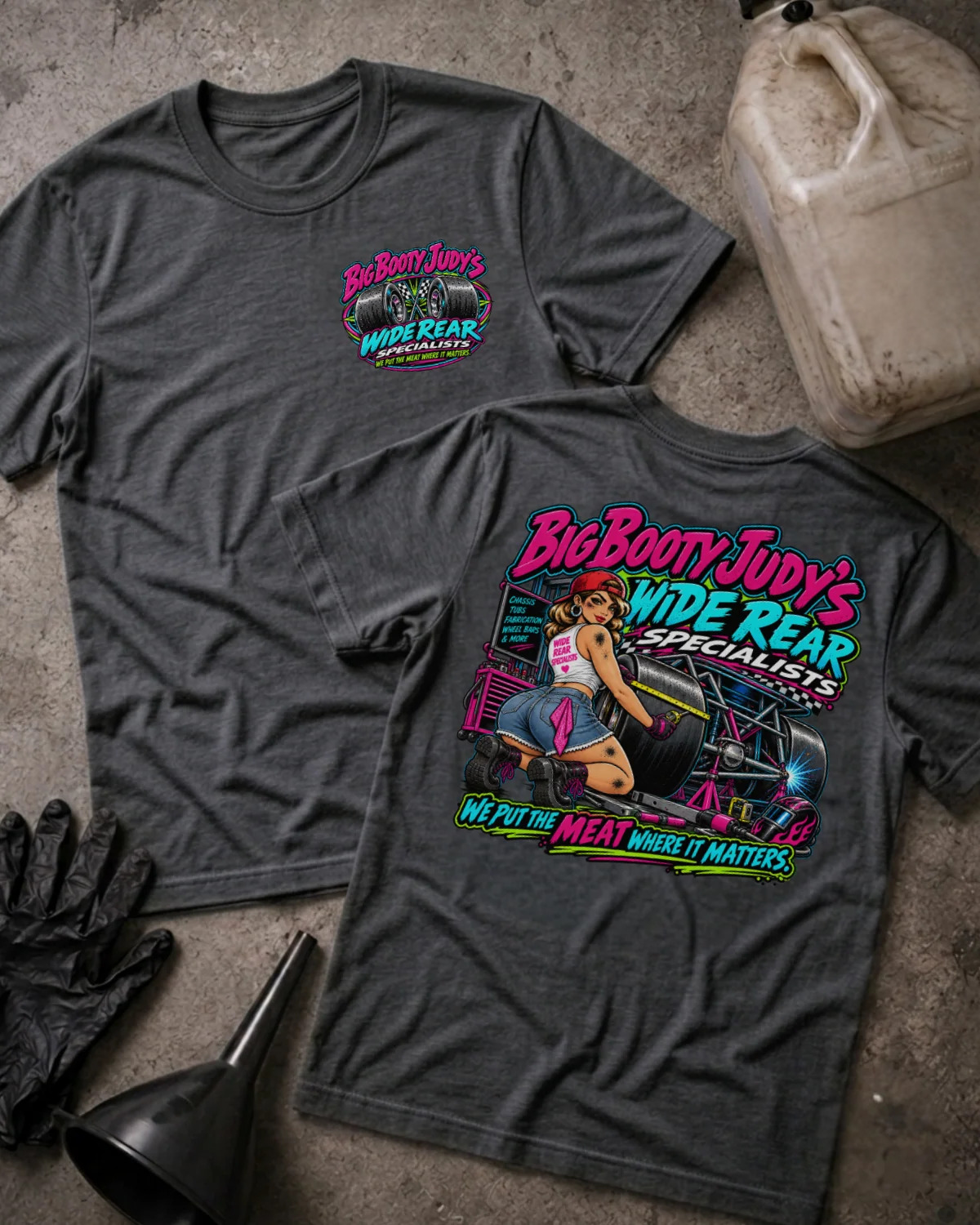 Two dark heather gray graphic T-shirts laid flat on a concrete-like surface, with a black funnel, gloves, and a plastic jug as styling props. The front shirt shows a small pink and teal chest graphic reading Big Booty Judy's Wide Rear Specialists.