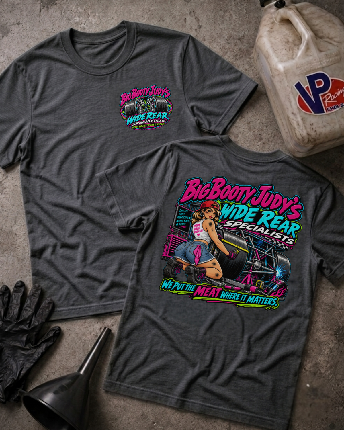 Two dark grey heather graphic T-shirts laid flat on a concrete surface, showing the front and back designs for 'Big Booty Judy's Wide Rear Specialists'. The front features a small pink and green logo on the left chest, while the back displays a large, colorful pin-up style graphic with a woman and the brand name.