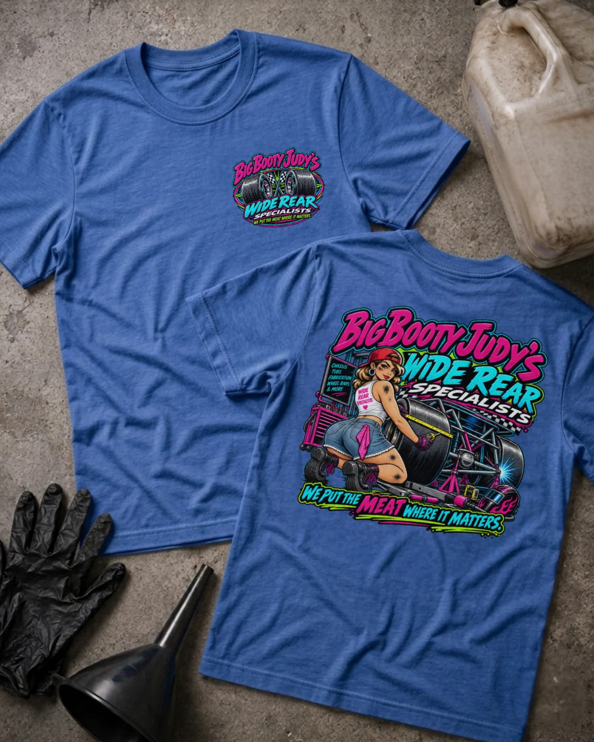 Two heather true royal graphic T-shirts laid flat on a concrete surface, showing the front and back designs. The shirts feature a colorful 'Big Booty Judy's Wide Rear Specialist' graphic with a pin-up illustration and hot rod elements